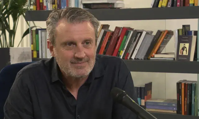 Uomo con barba grigia sorride, microfono davanti. Alle sue spalle scaffali pieni di libri, tra cui "Le ossa parlano trama".