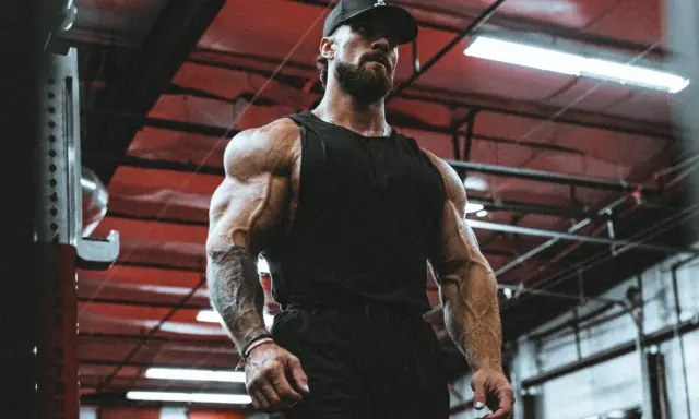Ein Mann mit beeindruckendem **mens physique** steht im Fitnessstudio. Seine Muskeln sind stark definiert, er trägt eine schwarze Kappe und ein Tanktop.