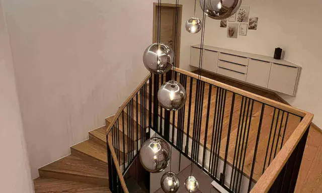 Nowoczesna klatka schodowa w domu z drewnianymi stopniami i czarną balustradą, ozdobiona wiszącymi, lustrzanymi kulami.