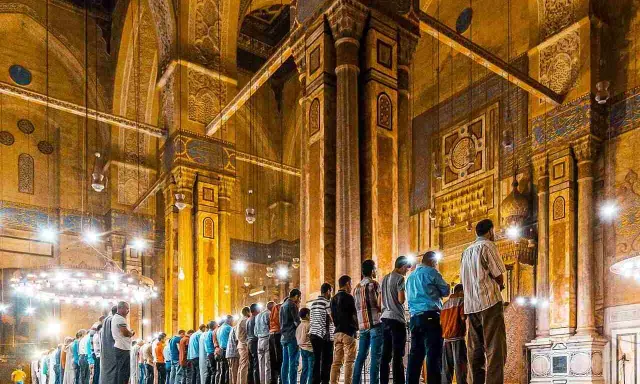 Männer beten Tarawih in einer Moschee. Sie stehen in Reihen vor kunstvoll verzierten Säulen und Bögen, beleuchtet von zahlreichen Lampen.
