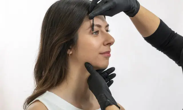Une esthéticienne ajuste les sourcils d'une femme avec une brosse, préparant un browlift pour un regard sublimé.