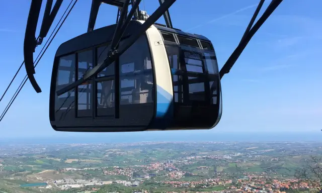 Cabina della funivia di San Marino in primo piano, con vista panoramica sulla città e sul mare all'orizzonte.