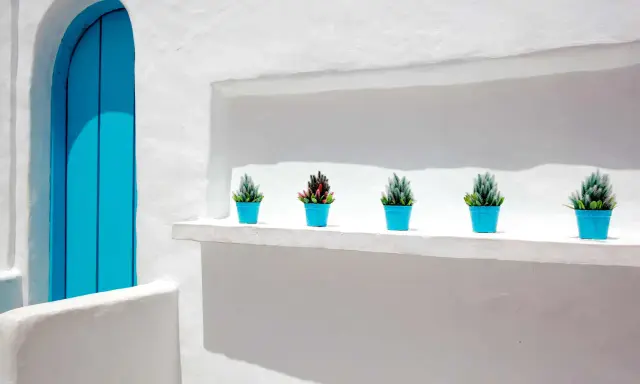 Fachada blanca con puerta azul brillante y cinco macetas con plantas. La textura de la pared sugiere que se usó pintar con cal.