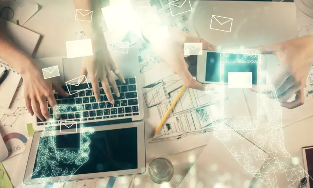 Dwie osoby pracują przy biurku, jedna pisze na laptopie, druga trzyma smartfon. Wokół latają ikony kopert, sugerując komunikację. Jak zacząć maila?