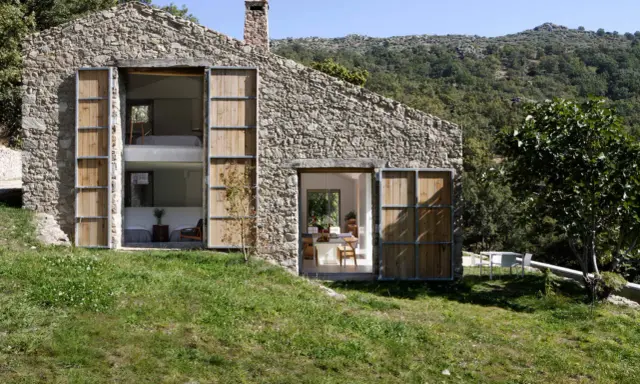 Una casa in pietra con grandi porte in legno, perfetta per chi ama vivere in campagna. Si intravedono interni moderni e un paesaggio collinare rigoglioso.