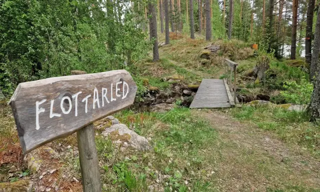 Träskylt med texten "Flottarleden Munkedal" pekar mot en liten träbro över en bäck i en skogsmiljö.