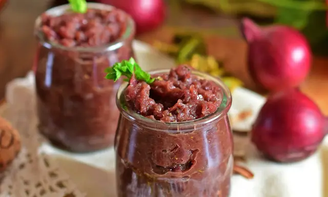 Dwa słoiczki z domowym, aromatycznym chutney z czerwonej cebuli, ozdobione listkiem zieleniny.