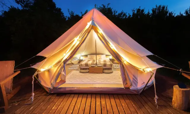 Mysig glamping i Sverige. Ett upplyst tält med säng och mysig inredning på en träaltan, omgivet av mörka träd.