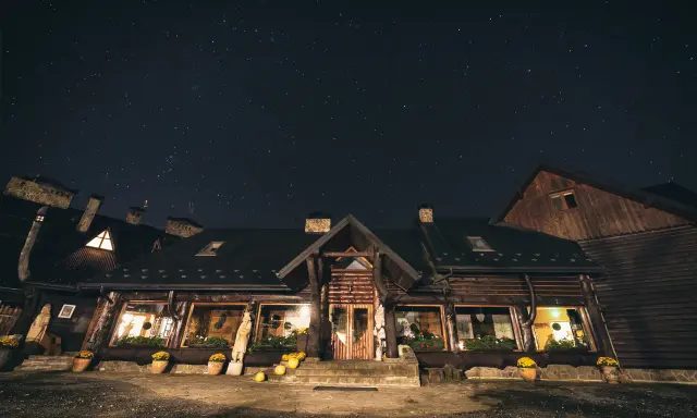 Nocne niebo nad bacówką Biały Jeleń. Drewniany budynek z oknami oświetlonymi ciepłym światłem, otoczony donicami z kwiatami.