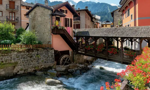 Un pittoresco ponte di legno attraversa un torrente impetuoso, fiancheggiato da case colorate e fiori rossi. Cosa vedere in questo incantevole borgo alpino.