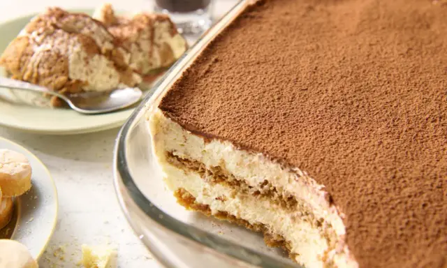 Pyszne tiramisu bez jajek, obficie posypane kakao, w szklanej formie. Obok porcja na talerzyku.