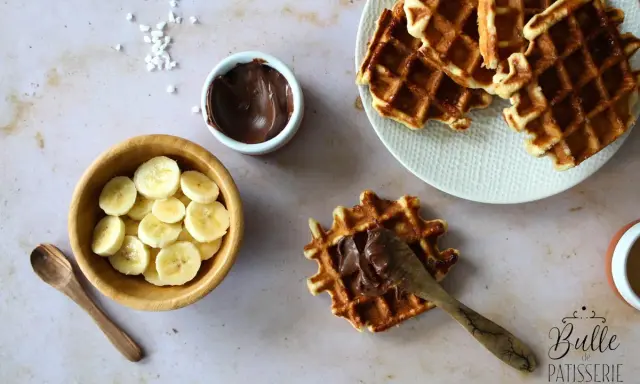 Une gaufre bubble est garnie de pâte à tartiner au chocolat. À côté, des rondelles de banane et une cuillère en bois.