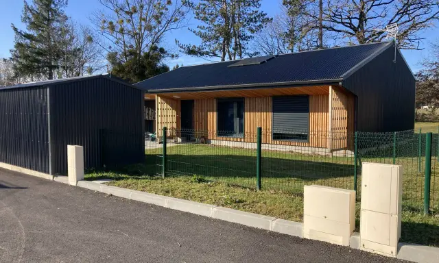Maison passive ossature bois avec panneaux solaires sur le toit, bardage bois et métal, devant un ciel bleu et des arbres.