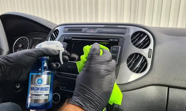 Ręce w czarnych rękawiczkach czyszczą deskę rozdzielczą samochodu, używając płynu "Wash Mist". To prosty sposób, jak pomalować plastiki w samochodzie, nadając im nowy wygląd.