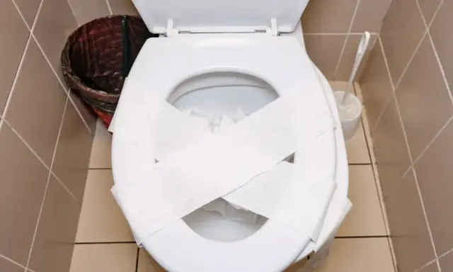 Inodoro blanco con papel higiénico cruzado sobre la tapa, listo para desatascar wc. Cerca, un cesto de basura y un cepillo.