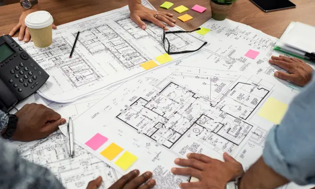 Des mains examinent un plan d'une maison, avec des post-it colorés et un téléphone sur la table.
