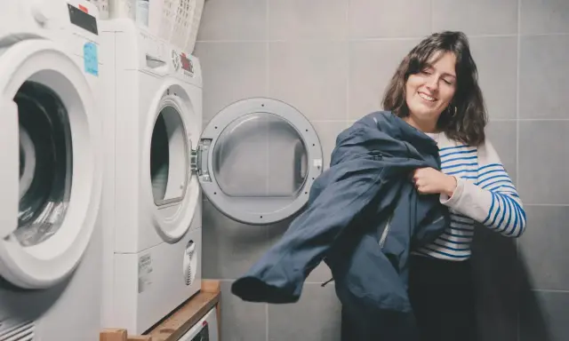 Kobieta z uśmiechem trzyma granatową kurtkę, przygotowując się do prania Gore-Tex. Obok stoi otwarta suszarka.