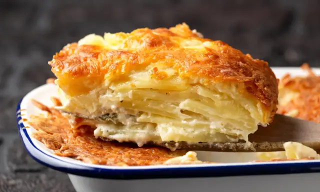 Une part généreuse de gratin de pomme de terre, avec sa croûte dorée et ses couches crémeuses, est soulevée par une spatule en bois.