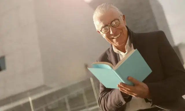 Ein älterer Mann mit Brille lächelt und hält ein hellblaues Buch. Er scheint Freude am Auswendiglernen von Gedichten zu haben.