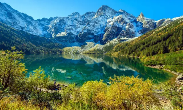 Widok na Morskie Oko z wysokości. Turkusowa tafla jeziora odbija ośnieżone szczyty Tatr. Jesienne drzewa otaczają brzeg.