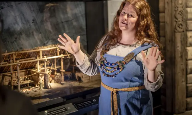En kvinna i vikingakläder berättar om en modell av en vikingagård på ett vikingamuseum.