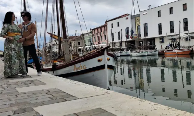 Coppia in attesa a Cesenatico, con barche d'epoca ormeggiate e il Municipio sullo sfondo. Un ricordo speciale prima di San Marino.