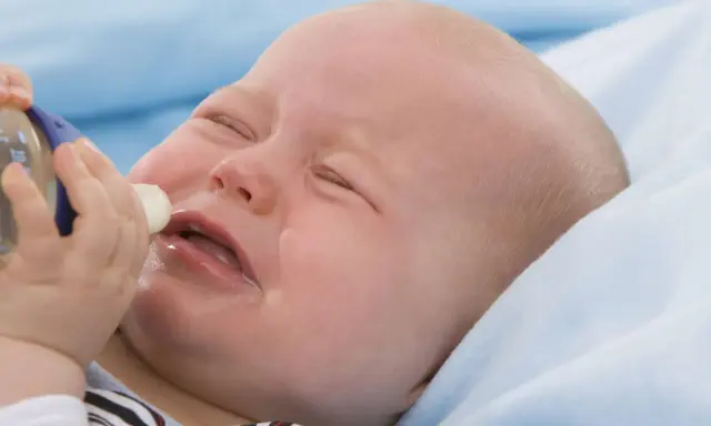 Ein weinendes Baby mit geschlossenen Augen, dem eine Flasche angeboten wird. Das Baby will nicht trinken.