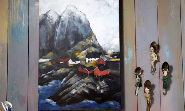 Målade landskap med röda hus vid en bergig kust. Konstverket hänger på en vägg av slitna träplankor.