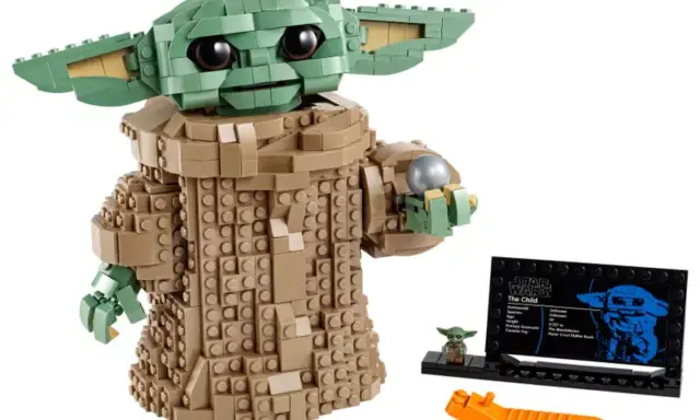 Figurka Grogu z klocków LEGO, znana z gier LEGO Star Wars, trzyma srebrną kulę. Obok stoi mała figurka i tabliczka informacyjna.