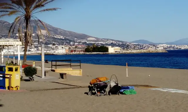 Sonniger fuengirola strand mit Palmen, Bergen und dem Meer im Hintergrund. Kinderwagen und Gepäck liegen am Sand.