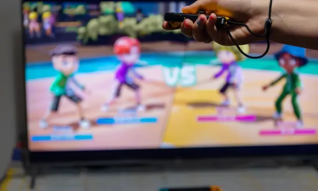 Ręka trzymająca kontroler do gry, na ekranie telewizora widać kolorowe postacie z gry ruchowej Nintendo Switch.