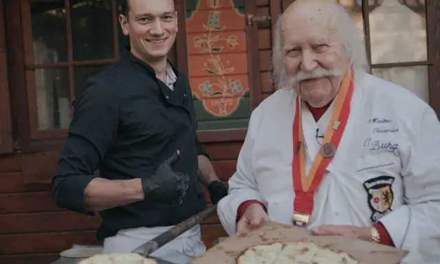 Deux chefs, l'un jeune et souriant, l'autre âgé avec une médaille, présentent des tartes flambées, évoquant l'origine du flamenkuch.