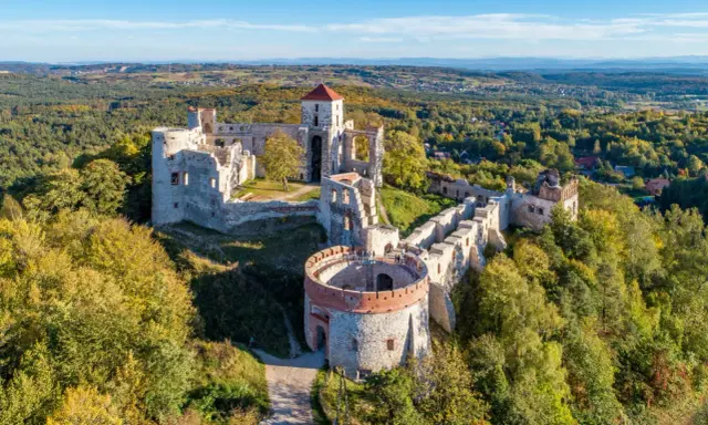 Ruiny zamku Tenczyn na wzgórzu, otoczone jesiennym lasem. Majestatyczne mury i wieże świadczą o dawnej potędze.