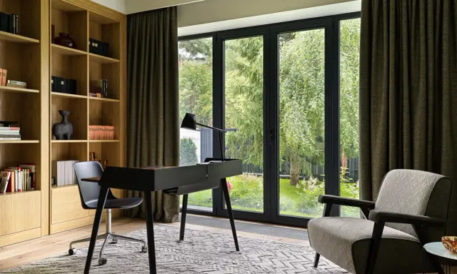 Acogedora habitacion despacho en casa con estanterías de madera, escritorio negro y sillón estampado. Ventanal con vistas al jardín.
