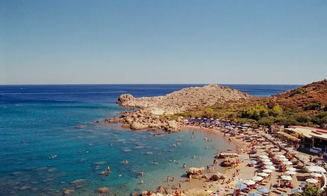 Rodos: turkusowe morze Egejskie, kamienista plaża z leżakami i parasolami, ludzie kąpiący się w wodzie.
