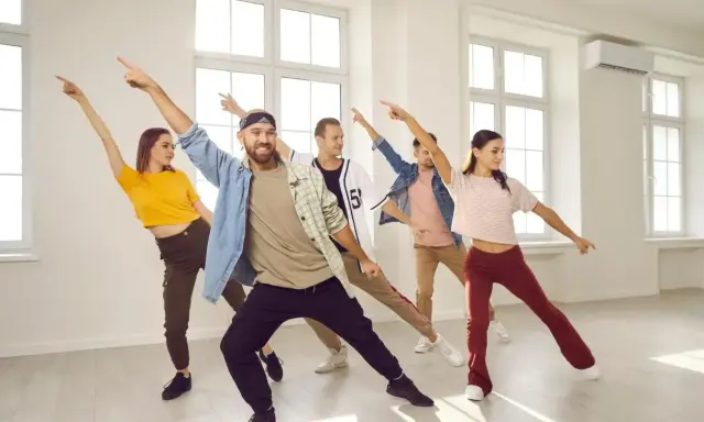 Grupa ludzi ćwiczy energiczne samba kroki w jasnej sali tanecznej, z rękami uniesionymi w rytm muzyki.