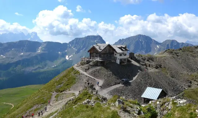 Rifugio alpino su un crinale roccioso, con sentiero tortuoso e escursionisti. Un'auto si avvicina al passo stalle.