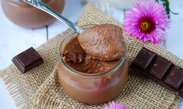 Une cuillère de crème au chocolat sans sucre, onctueuse et aérienne, sur un pot en verre. Des carrés de chocolat et des fleurs roses décorent la scène.