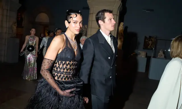 Dua Lipa stuns at the Met Gala in a black feathered gown and diamond necklace, walking with a companion.