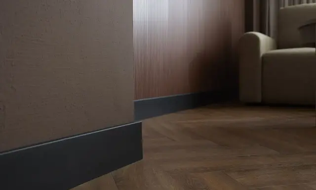 Ciemne listwy przypodłogowe przy brązowej i drewnianej ścianie, na tle parkietu i kanapy.