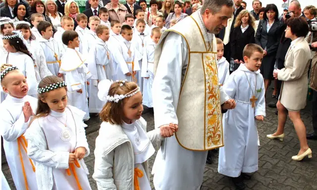 Dzieci w białych strojach, kiedy jest komunia, idą z księdzem.