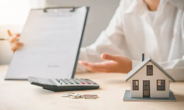 Une personne présente un contrat, une calculatrice, des clés et une maquette de maison. L'ambiance est un peu sinistre, comme si un problème était sur le point d'être révélé.