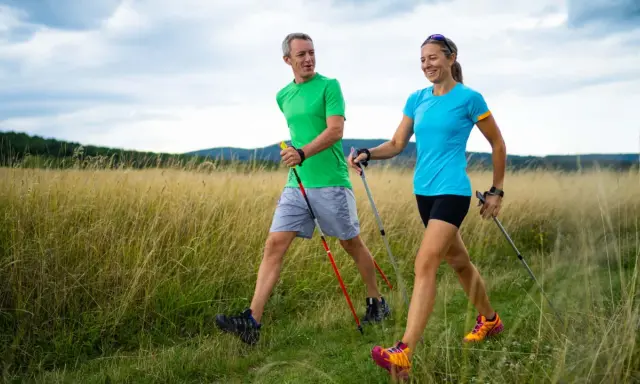 Ein Paar genießt das **power walking** in einer sonnigen Landschaft mit hohem Gras und bewaldeten Hügeln im Hintergrund.