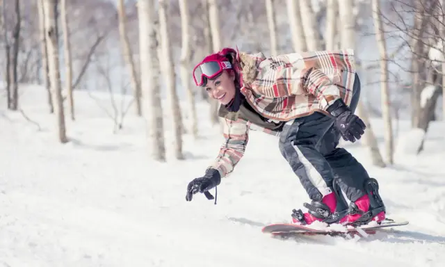 Uśmiechnięta snowboardzistka z różowymi włosami zjeżdża po ośnieżonym stoku wśród drzew. To świetna zabawa podczas chodzenia po górach zimą.