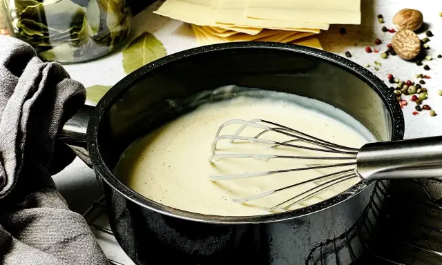 Cremige Béchamelsauce wird in einem Topf mit einem Schneebesen zubereitet. Daneben liegen Lasagneplatten und Gewürze.