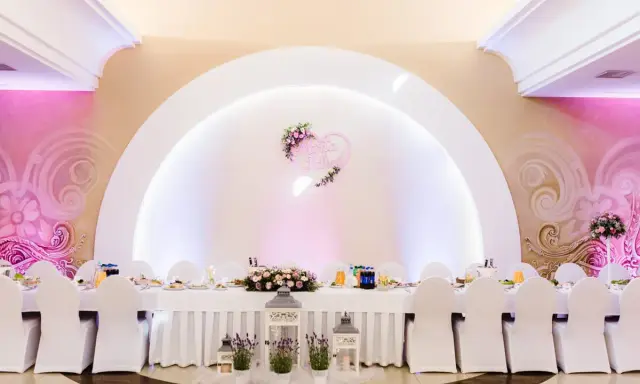 Elegancka sala weselna z białymi pokrowcami na krzesłach i serwowanym stołem. Serce z imionami pary młodej zdobi ścianę, tworząc atmosferę wesela z klasą.
