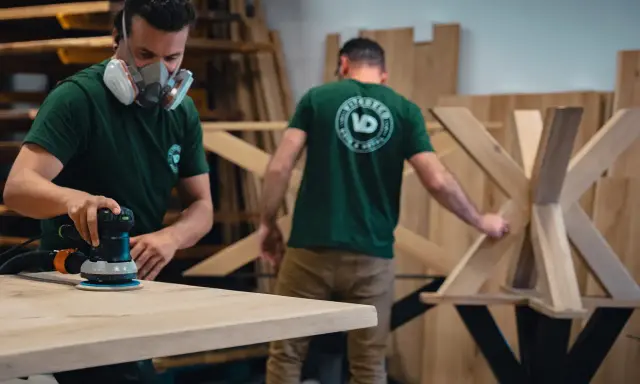 Un artisan porte un masque et utilise une ponceuse pour poncer une table en bois. Un autre artisan travaille sur un pied de table en bois.
