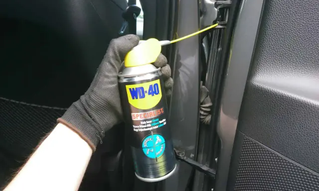 Ręka w rękawiczce aplikuje biały smar litowy WD-40 na zawiasy drzwi samochodu, by je nasmarować.