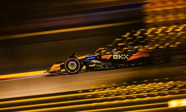 Un coche de **fórmula 1** naranja y negro acelera en la noche, dejando estelas de luz dorada.