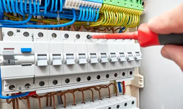 Main électrique avec disjoncteurs. Un tournevis rouge est utilisé pour ajuster une connexion, aidant à comprendre comment savoir quel fusible changer.